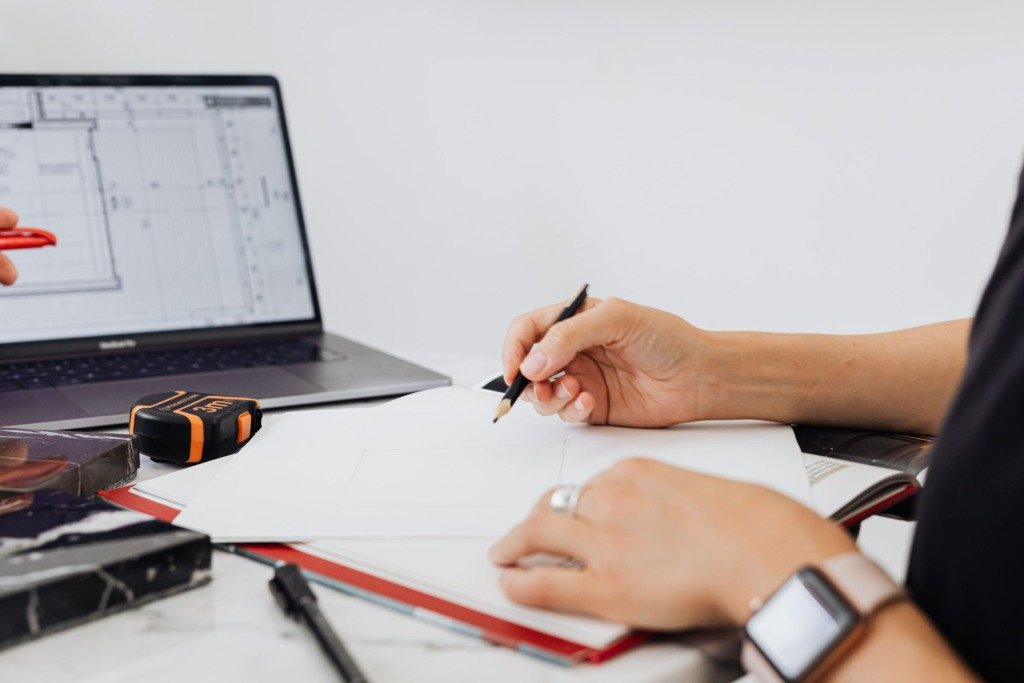 Close-up of an interior designer sketching plans with a laptop displaying architectural designs.